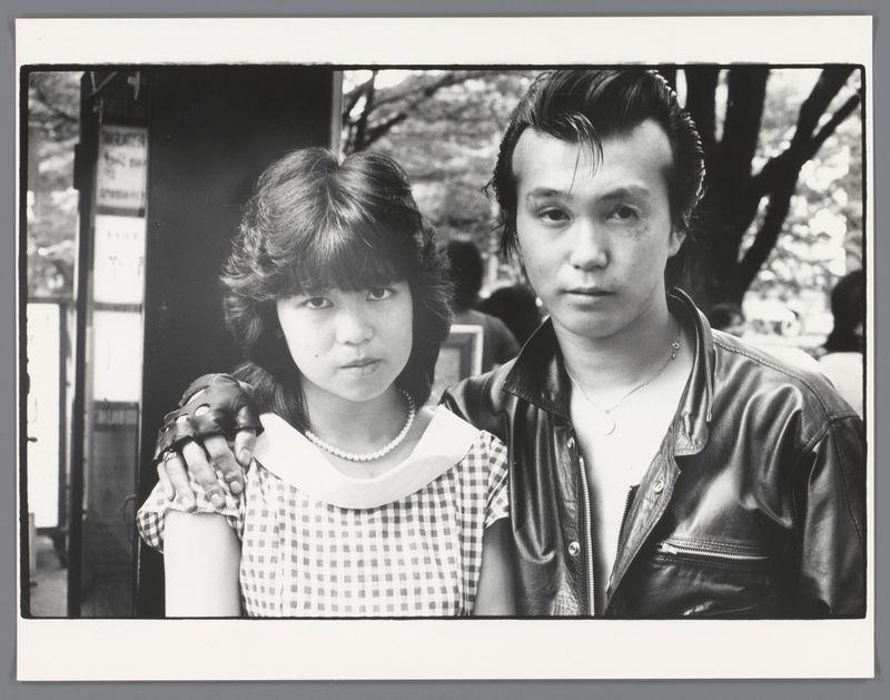 The image is a black and white photograph featuring two individuals, likely taken during the daytime outdoors. On the left side of the photo stands an individual with shoulder-length hair styled in waves or curls, wearing what appears to be a sleeveless checkered dress with a collar, accessorized by a necklace that features circular beads. Their hands are clasped together over their stomach area.

On the right is another person dressed in a leather jacket and sporting a hairstyle reminiscent of early rock 'n' roll styles from the mid-20th century. This individual wears minimal jewelry—a simple pendant around their neck—and has visible tattoos on both arms, suggesting an interest or involvement in tattoo culture.

The background suggests they are at an outdoor location with trees indicating it might be during spring or summer based on leaf presence and sunlight filtering through branches. A signpost is partially visible behind them, but the text cannot be discerned from this angle. 