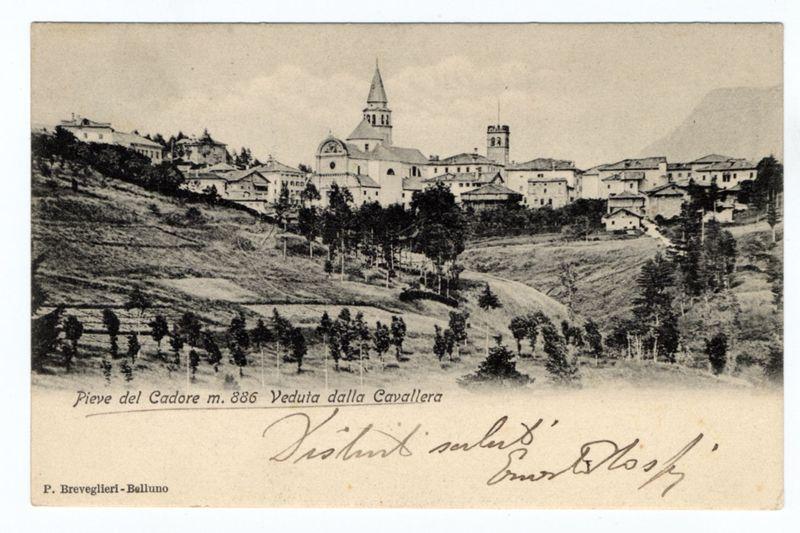 the image is a black and white photograph depicting an elevated, sprawling village nestled on the side of a hill. The architecture suggests European influences with several buildings featuring pitched roofs and what appears to be traditional construction materials such as stone or wood. A prominent church-like structure stands out in the center of this community, showcasing pointed arches above its doors and windows and a spire reaching towards the sky.

The terrain is predominantly forested at lower elevations which gradually transitions into open fields higher up on the hillside. The landscape seems tranquil with no visible modern vehicles or infrastructure that would indicate contemporary time periods beyond what could be inferred from architectural styles.

Inscriptions within the image include "Pieve del Cadore m. 886 Veduta dalla Cavalleria" and a handwritten note in cursive script, which translates to 'Village of Cadore at an altitude of 886 meters Viewed by Cavalry' followe