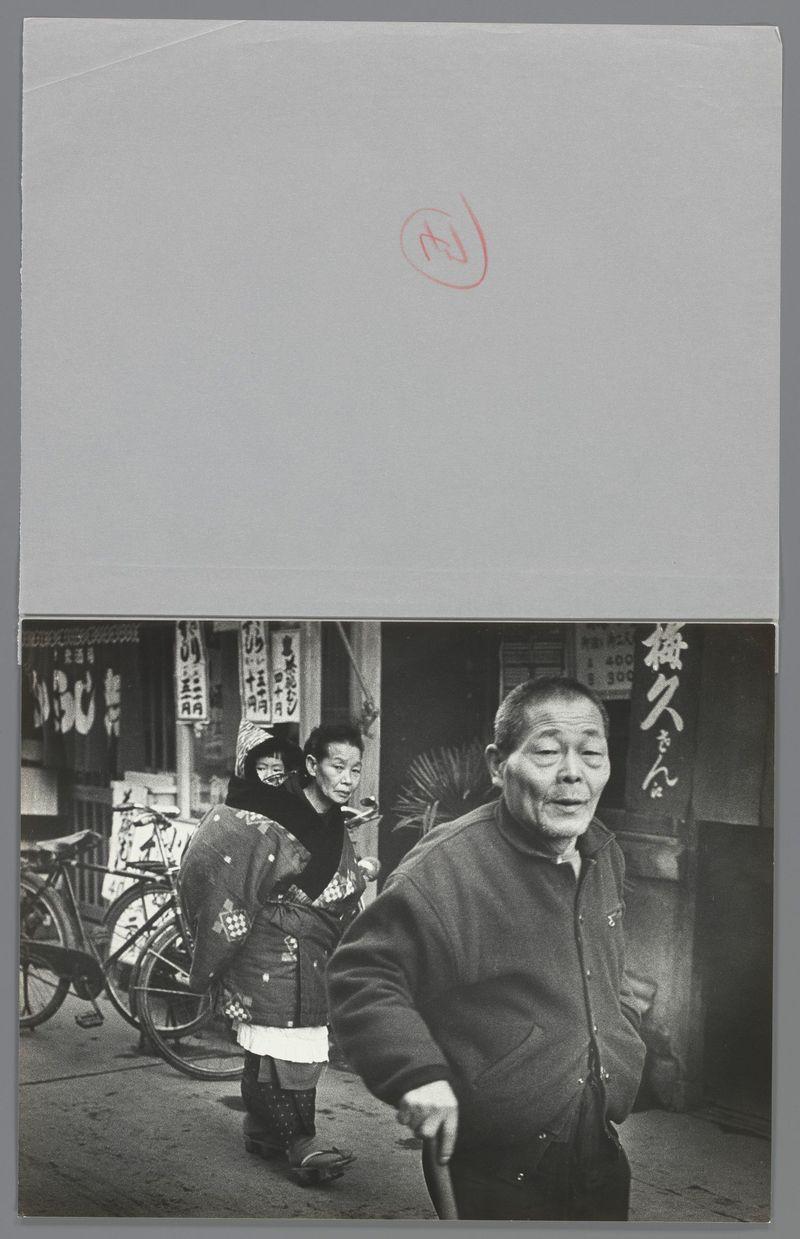 The image depicts a candid street scene in Japan from the late 1950s or early 1960s, captured by photographer Ed van der Elsken. It features three individuals: an elderly man wearing glasses and traditional attire with floral patterns is walking forward, holding his cane; behind him stands another person who appears to be a child in school uniform, looking at something out of frame. In the background, there's a third individual seen from behind, engaged in conversation or reading material near several bicycles parked against storefronts.

The black and white composition conveys an atmosphere reflective of daily life during that period with visible signage written in Japanese characters indicating businesses such as a vegetable shop and other local establishments. The image is printed on glossy paper, possibly part of a publication or exhibition print from the photographer's archive, indicated by the red circle with '83' handwritten at the top right corner.

The photograph captures 