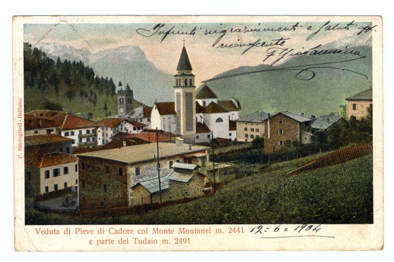 The image is a vintage postcard depicting a scenic view of the Pieve di Cadore village, specifically highlighting Monte Montanel and part of Tudaio. The scene shows traditional Italian architecture with buildings featuring red-tiled roofs and whitewashed walls. A prominent clock tower stands in the center against a backdrop of gentle hills adorned with greenery and trees under an overcast sky. Handwritten annotations on the right side include what appears to be names, possibly referring to individuals associated with or interested in this location. Below these inscriptions is printed text providing information about the buildings shown: "Veduta di Pieve di Cadore col Monte Montanel m. 2441" and "parte del Tudaio m. 2491," suggesting measurements related to elevation above sea level. In red, there's a handwritten note with calculations likely indicating years or dates associated with the location. The overall image has signs of wear consistent with an antique postcard.