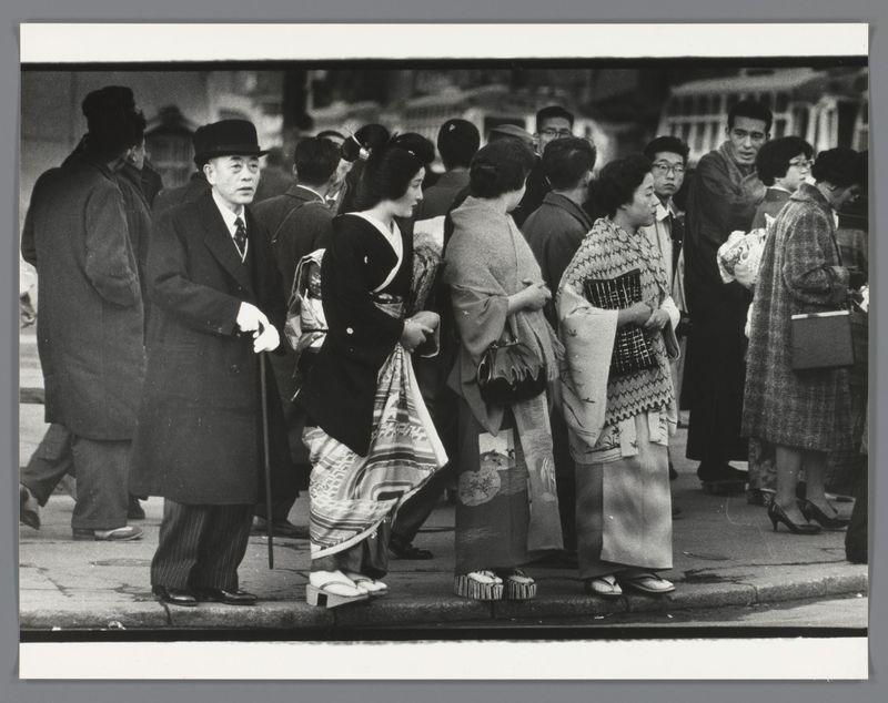 The image depicts a bustling street scene with numerous individuals gathered. The foreground shows people standing and conversing, wearing varied attire indicative of different social statuses or roles. On the left side stands a man in a dark coat and hat, holding onto his jacket as he engages with others around him. In contrast to this formal style, several women display traditional Japanese clothing such as kimonos adorned with intricate patterns and elaborate designs. These garments include layers called obi belts which add to their ornate appearance.

The crowd is densely packed on the sidewalk, suggesting a popular or significant area within Japan during that period. The individuals' expressions vary from engaged in conversation to passive observation of their surroundings. Notably, some women carry handbags and boxes indicative of shopping activities typical for a market-like setting. The image has a historical feel due to its black-and-white composition, reflecting the fashion
