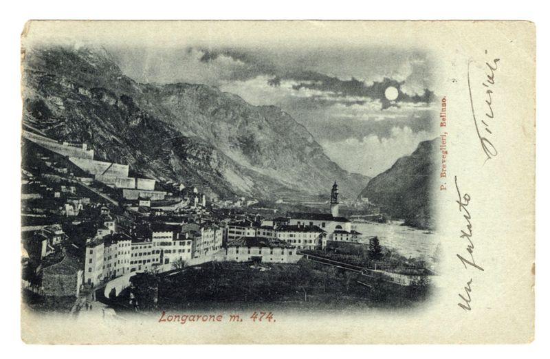 The image is a black and white postcard depicting an old town situated in a mountainous region. The village appears to be nestled along the side of a river, with buildings densely packed together on steep slopes leading down towards the water's edge. Prominent features include several large structures that resemble churches or municipal buildings, particularly one at the uppermost part which has a tower and what looks like an observation deck. In the background, imposing mountains rise dramatically from either side of the valley where the village is located.

The sky above shows some clouds with a distinct circular object resembling the moon near the top right corner, suggesting it could be captured during twilight or nightfall when this image was taken. The postcard bears handwritten and printed inscriptions indicating its origin as "Longarone m. 474" in Belluno Province of Veneto region.

Additional notes on the card include a cursive signature that appears to read 'Ferrovia,' po