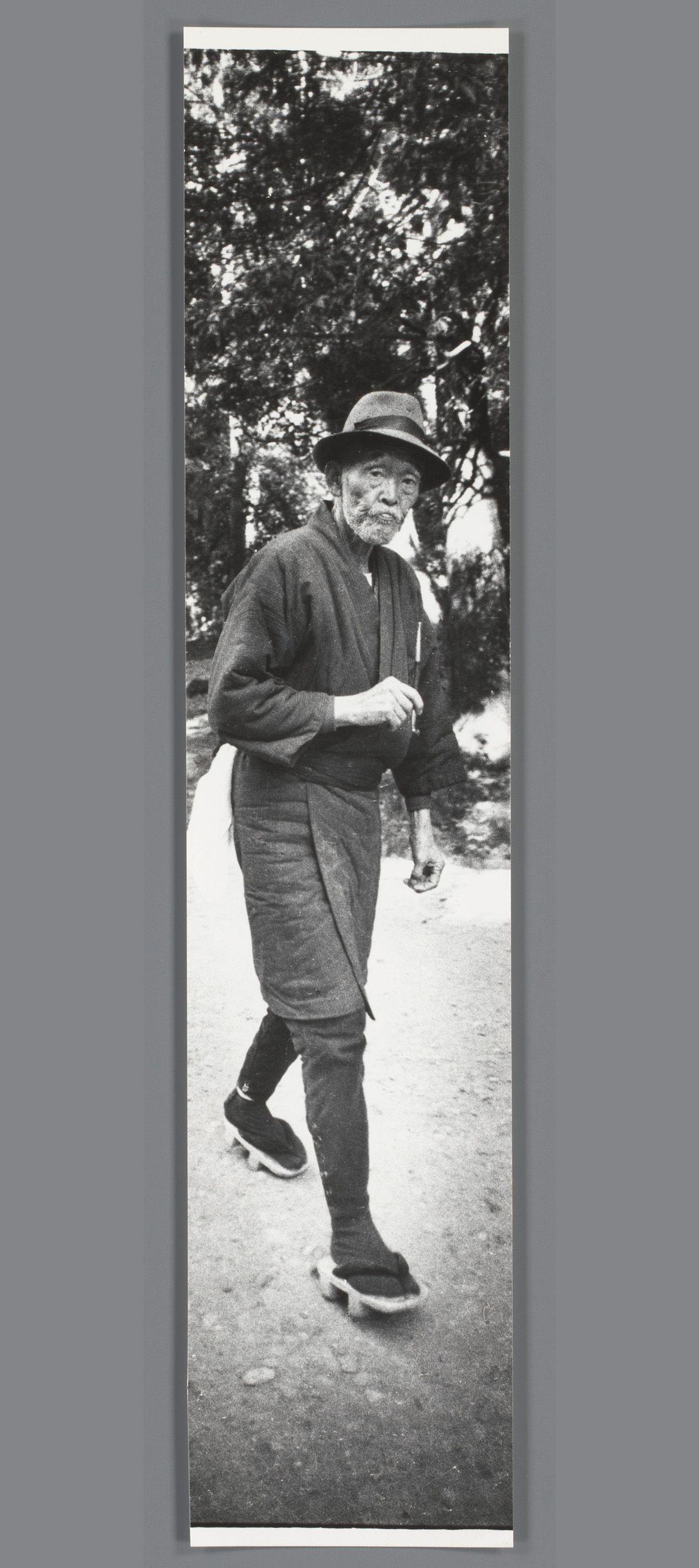 The image is a black and white vertical portrait photograph of an elderly man walking on what appears to be a path with trees in the background. The subject, who looks contemplative or introspective, has a distinctive beard and mustache, which frames his face as he gazes slightly downwards. He wears traditional attire that includes a long-sleeved robe-like garment (which could indicate cultural significance), dark trousers, heavy-duty shoes for walking on uneven terrain, possibly indicating rural settings, and is holding something in one hand while the other hangs by his side.

The photograph has an overall grainy texture indicative of older film photography. The man's posture suggests a steady pace as he walks, with visible movement captured through slight blur. This could signify either actual motion or be a result of camera technique to convey dynamism within the stillness of the portrait.