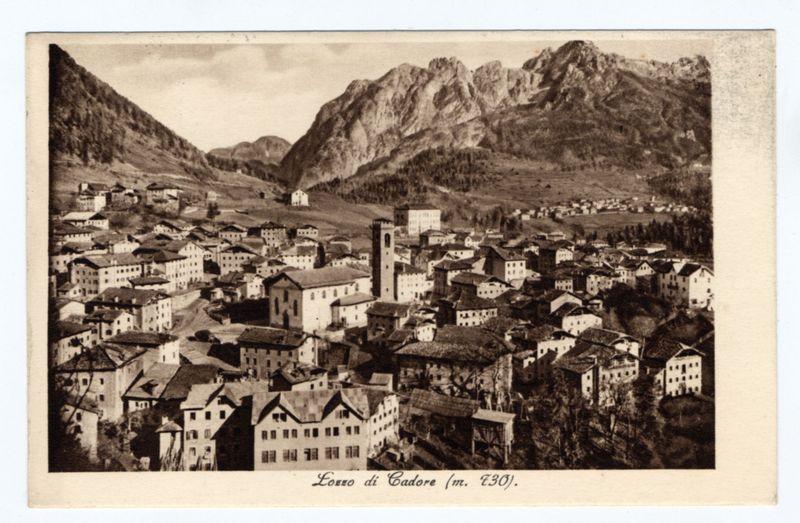 The image depicts a sepia-toned photograph of the town of Lozzo di Cadore, dating back to around 1930 as indicated by the inscription. It is presented in portrait orientation and shows an elevated view overlooking the densely built-up area surrounded by mountainous terrain with prominent peaks visible in the background.

In this historic scene, a variety of buildings are seen clustered together on terraced slopes indicative of traditional hillside settlements typical for regions within or near mountain ranges. Prominent among these is a tall structure with what appears to be a tower and dome at its peak, possibly indicating religious significance like a church or bell tower. The architecture suggests European influence, likely from the Italian alpine region.

The photograph's monochromatic coloration gives it an antique feel, suggesting that it has been preserved over many decades since its creation. There is text on the image which reads "Lozzo di Cadore (m. 130)," indicating a sp