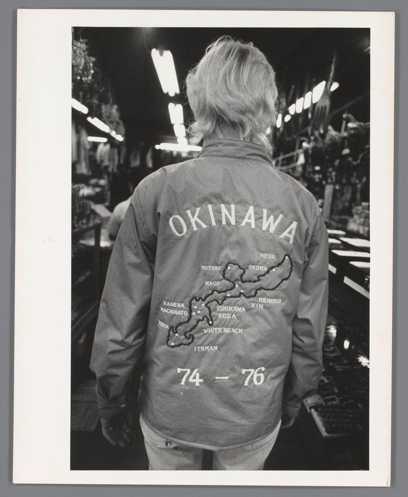The image is a black and white photograph showing the back of an individual wearing a grey jacket with text and graphics on it. The jacket displays "OKINAWA" prominently at the top, followed by what appears to be a map outline of Okinawa Island below which lists several places such as Naha, Motobu, and others in Japanese characters. Below this list is more English text: "HEDO," "MOTOBU," "OKUMA," etc., also corresponding with various locations on the island's map. There are additional words like "NAGO" and numbers "74-76." The bottom right of the jacket has a small figure resembling an Okinawan person, labeled as "ITOMAN." At the very bottom center, it states "1974 - 1976," suggesting this garment is commemorative or indicative of service during those years. The background shows industrial shelving with lights on display shelves, hinting at a warehouse-like setting.
