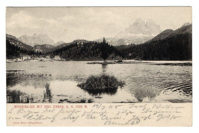 The image depicts a vintage postcard featuring an old black and white photograph. The scene captures Misurina-See, also known as Lake Misurina with the Three Peaks of Lavaredo in the background. This picturesque lake is surrounded by dense forested hills leading up to majestic mountains. A small island or landmass can be seen rising from the center of the water body. In the distance on one side of the lake sits a large building, likely an inn or hotel, with several smaller structures around it indicating some form of habitation or tourist facilities.

The sky is overcast, giving the photograph a somewhat somber and nostalgic feel. Handwritten text at the bottom left corner provides additional context: "MISURINA-SEE MIT DREI ZINNEN, S. H. IS0O M., 19/7 '65" suggests it was taken on July 24th in the year of 1865 and is signed by Alois Beer from Klagenfurt.

The bottom right corner contains more handwriting which appears to be a personal note or message, perhaps added later. The overa