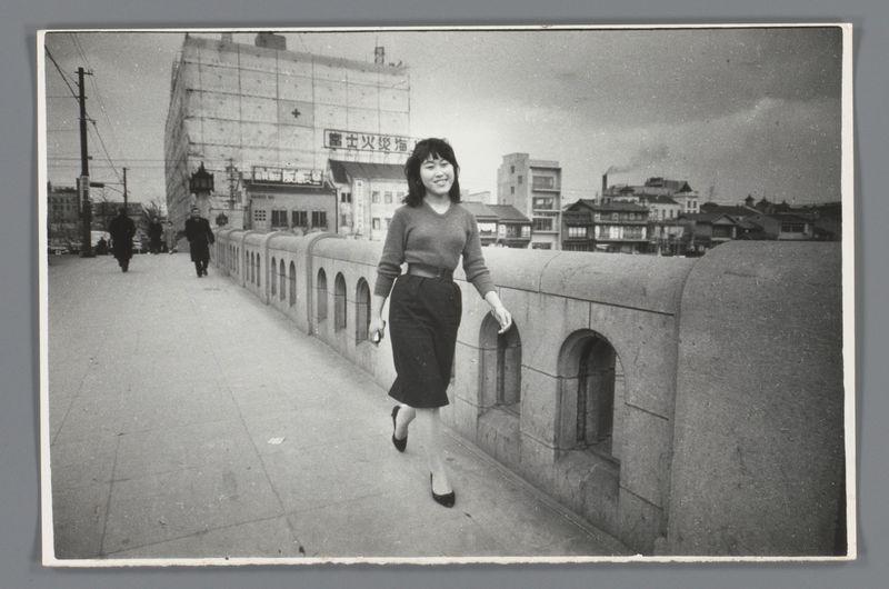 The image is a black and white photograph depicting an East Asian woman walking on what appears to be a pedestrian bridge. She has shoulder-length hair, wears glasses perched atop her head, a long-sleeved grey top, a dark skirt with high-waistline cinched at the waist by a belt or sash, and closed-toe black heels. Her posture is relaxed as she strides along the pavement which runs parallel to another walkway occupied by two distant figures in dark coats.

The setting suggests an urban environment during mid-20th century Japan; this inference comes from several clues: architectural styles of buildings with signs bearing non-Latin characters, likely Japanese script and language. The architecture includes a mix of traditional wooden structures indicative of older districts alongside more modern concrete block designs. A significant building in the background appears to be under construction or renovation given its unfinished facade.

The sky is overcast, suggesting either early mornin