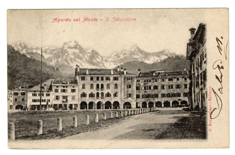 The image shows a vintage black and white photograph of an old building with multiple floors, featuring rows of arched windows and balconies. The structure has a prominent central section flanked by wings on either side. In the background, snow-capped mountains rise against a pale sky, suggesting that this location is in or near a mountainous region during winter.

In red cursive text at the top left corner, it reads "Agordo col Monte - S. Sebastiano," indicating the name of the place and its geographical feature. A handwritten signature on the bottom right says "Guglielmo M.", likely referring to Wilhelm Mahler, a famous photographer known for his work in Turin during that era.

The image also includes two lines of text at different orientations: one running along the top edge with red lettering saying "Agordo col Monte - S. Sebastiano," and another on the bottom right corner with cursive writing attributing it to P. Breverlièri-Belluno, possibly a photographer or creator associat