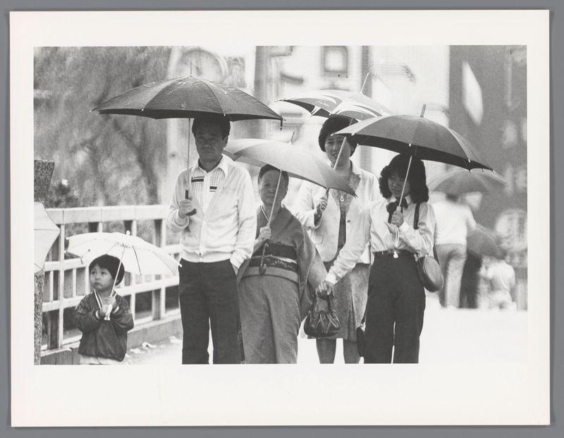 The image is a black and white photograph depicting multiple individuals walking on what appears to be a rainy day. The focus of the photo captures several people holding open umbrellas, shielding themselves from rain or possibly snow. In the foreground, there are four adults carrying dark-colored umbrellas; two men in suits with ties and one woman dressed more casually. Behind them is another person partially visible under an umbrella. On the left side, a child wearing traditional Japanese attire and shoes can be seen holding onto something small and white while walking alongside an adult also under an umbrella.

The background is blurred but shows signs of urban life: buildings or structures with windows are faintly discernible behind the people. The image has a candid quality to it as if capturing a spontaneous moment on city streets during inclement weather. There's no clear indication of specific locations within Japan, nor does there appear to be any explicit context about why 
