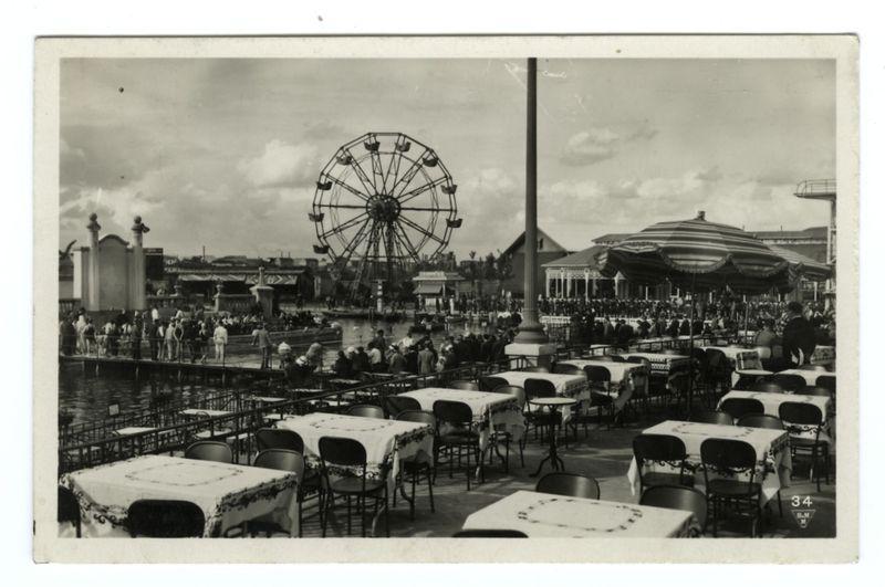 The image depicts a vintage black and white photograph of an amusement park area. In the foreground, there's an outdoor dining area with multiple tables covered in cloth tablecloths featuring floral patterns, surrounded by chairs. The atmosphere suggests it could be early to mid-20th century based on the style of clothing worn by patrons and furniture design.

In the middle ground, a Ferris wheel stands prominently against the sky, indicating the presence of an amusement park or fairground nearby. Beyond this, there's what appears to be water with people walking along its edge. The background is filled with various structures that could house rides or games typical for such parks, and several buildings are visible.

The composition suggests a lively yet relaxed public space designed for leisure activities. There’s also text in the image indicating "34 B.M", which might refer to an album number from Berliner Moden (Berliner Mode), as well as other indistinct markings that could be r