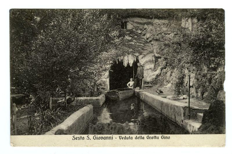 The image depicts a vintage black and white photograph of an entrance to the "Grotta Gino," which is likely located in Sesto San Giovanni. The photo shows a man standing at the edge of a cave-like opening, holding what appears to be a small boat or canoe above water that leads into the grotto. Vines are visible cascading down the rocky sides leading up to and within this entrance, suggesting it's nestled among vegetation. A pathway can be seen leading towards the entryway with steps descending toward the water level where the boat is placed for possible use as a means of exploration or transportation through the cavernous space ahead.

The image has text at the bottom that reads "Sesto S. Giovanni - Veduta della Grotta Gino," indicating it's from Sezano, San Giovanni and refers to a view (veduta) of this grotto named after 'Gino'. This description aims to provide an accurate representation for those who cannot see the image while emphasizing its historical context and location.

Th