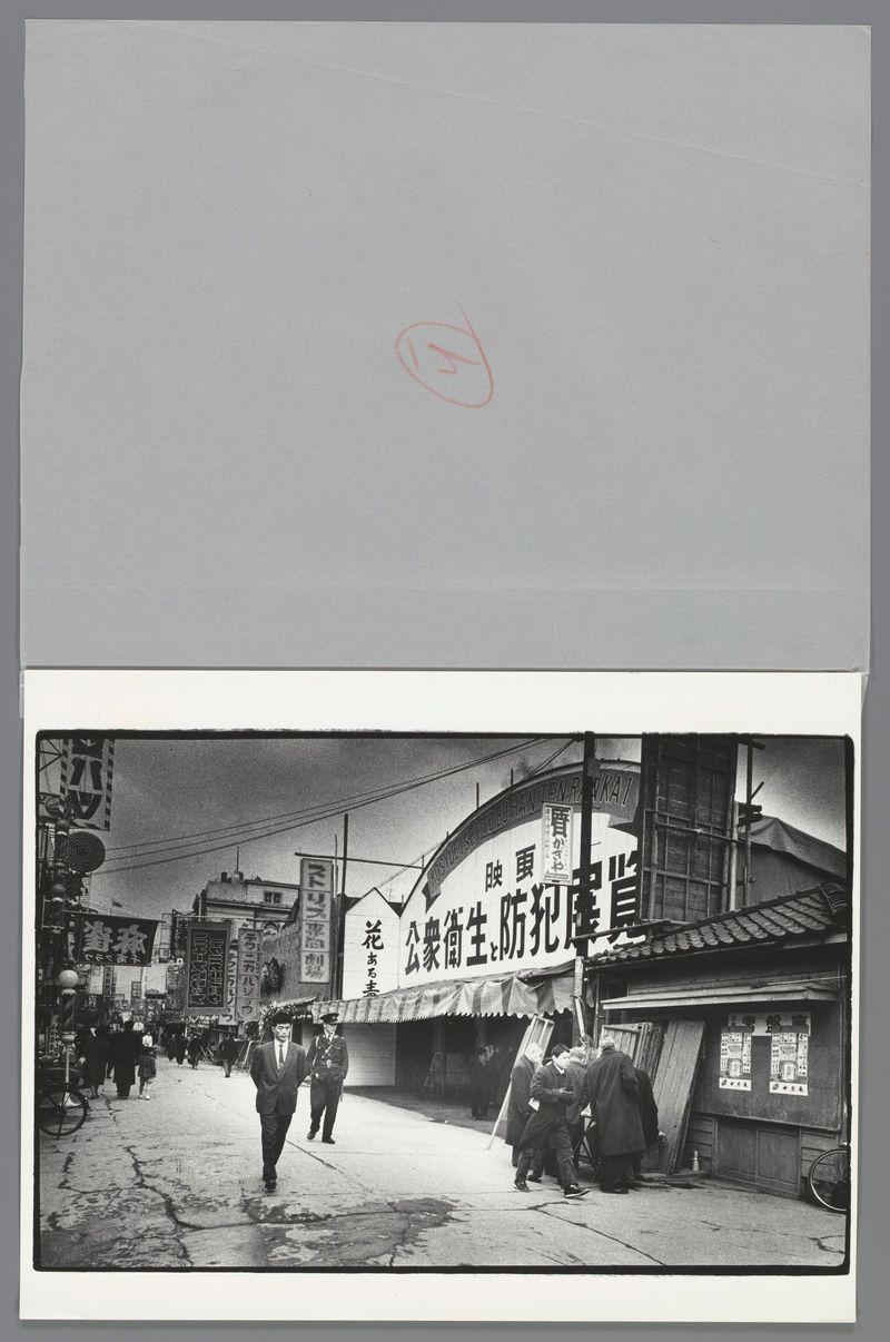 The image depicts a bustling street scene with multiple people walking and interacting in what appears to be an Asian urban environment, possibly Japan given the context provided. Prominently featured is signage for various establishments including restaurants or bars, indicated by Japanese characters on banners and awnings. The architecture has traditional elements mixed with more modern styles, suggesting this may be a historical district undergoing some degree of revitalization.

The black-and-white tone captures details such as texture, shadows, and contrast vividly, highlighting the busy atmosphere with groups of people in various states of activity—some walking solo or together, others engaging near storefronts. Notably, there is no direct indication of specific brands or services due to language barriers; however, the overall impression suggests a snapshot from daily life within this cultural setting.

The photograph carries an air of candid spontaneity and documentary style