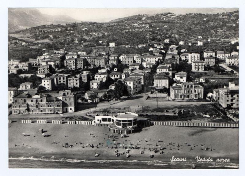 The photograph is a black and white aerial view of the coastal town of Scampia, with various buildings spread across the landscape. The foreground features an expansive sandy beach dotted with numerous sun loungers and umbrellas, indicating recreational use by visitors or residents. A prominent building complex near the water's edge appears to be a hotel or public facility, surrounded by smaller structures that could serve as additional accommodation or amenities for tourists.

In the mid-ground, more densely populated residential areas are visible, showcasing multi-story buildings with flat roofs and varying architectural styles. The background reveals rolling hills with vegetation covering some parts of it, which suggests a semi-rural setting beyond the urbanized area. The topography indicates that Scampia is situated in an area where coastal development meets hilly terrain.

The photograph captures what seems to be a typical sunny day at the beach and within the town, likely aim