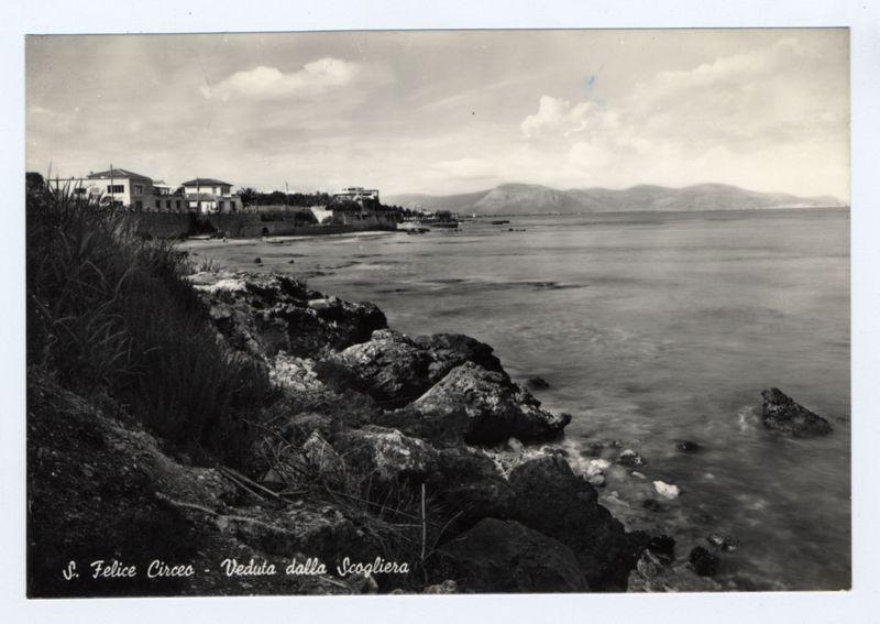 The image depicts a black and white photograph of a coastal scene. It captures rocky shores in the foreground leading into calm waters with gentle waves lapping against them, extending to an expansive view towards landforms in the distance.

In the midground lies a series of buildings on elevated ground adjacent to the shore, possibly residential or part of a small town. There's notable contrast between the man-made structures and the natural landscape around it.
Further back across the water, mountains are visible under a partly cloudy sky suggesting varied topography beyond what is immediately apparent from this perspective.

The photograph includes text at its bottom which reads "S. Felice Circe - Veduta dalla Scogliera", indicating that the location of the photo is S.Felice Circe and it was taken 'From the Rock'. There are signs of wear or damage to the edges, as well as discoloration on some parts of this physical photograph, which may affect its overall condition.