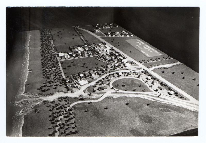 The image depicts a detailed black and white aerial view of what appears to be an early 20th-century coastal town. The model showcases various buildings, streets, fields, and green spaces. On the left side of the composition is a beach with waves crashing against it; rocks are scattered along its edge.

In the center lies a small cluster of houses surrounded by open land, which could represent agricultural plots or undeveloped areas. To the right, there's an expansive field labeled "Contro Riservata" (Italian for Restricted Area), indicating this part might have been used as military grounds at some point in history.

The model includes roads leading to and from these buildings, with vehicles parked along a main road that runs through the town. There is also a prominent curved area resembling an amphitheater or sports field within the green space near "Contro Riservata."

Overall, this image serves as a historical snapshot of urban planning and land use in early 20th-century Ital