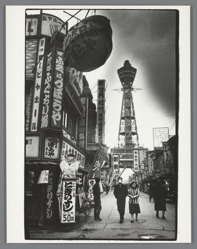 The image is a black and white photograph depicting an urban street scene in Japan, likely from the late 1950s or early 1960s given its vintage appearance. The setting appears to be a bustling commercial area with various signs written in Japanese characters indicating shops such as restaurants, bars, and other businesses. A large Ferris wheel stands prominently on the right side of the photo, adding an element of amusement park-like charm to the urban landscape.

Several people are visible walking down the street, dressed in what looks like mid-20th-century attire for Japan's citizens at that time: men wearing suits or casual coats and women wearing dresses with striped patterns. The expressions on their faces suggest a sense of everyday activity without any significant interaction captured between them. The architecture features tall buildings with numerous windows reflecting the overcast sky, suggesting an urban environment.

The photograph is credited to Ed van der Elsken, as i