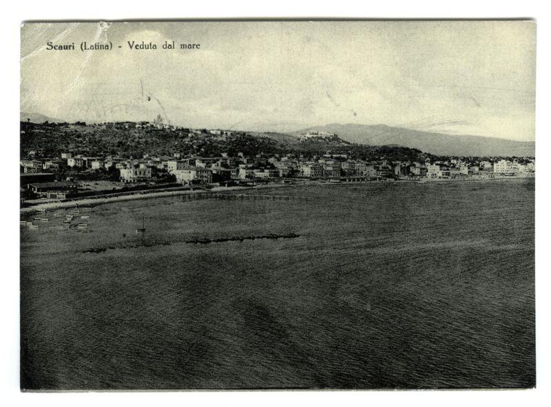 The image depicts a black and white photograph of the coastal town of Scuari, also known as Latina. The view is taken from the sea looking towards the shore with multiple low-rise buildings densely packed along the coastline on an elevated plain. In the distance are rolling hills or mountains partially obscured by atmospheric haze, suggesting a possible misty environment at the time the photo was captured.

In the foreground of the image lies calm waters that extend to meet the land near what appears to be a small harbor area with several boats docked along the shore and some floating in the water. The sky is overcast, adding to the monochromatic aesthetic typical for photographs from earlier periods when black-and-white film was prevalent.

The photograph's caption reads "Scuari (Latina) - Veduta dal mare," which translates to "Scuari (Latina) - View from the sea" in English. The text indicates that this image is a historical snapshot, likely dating back several decades or even ce