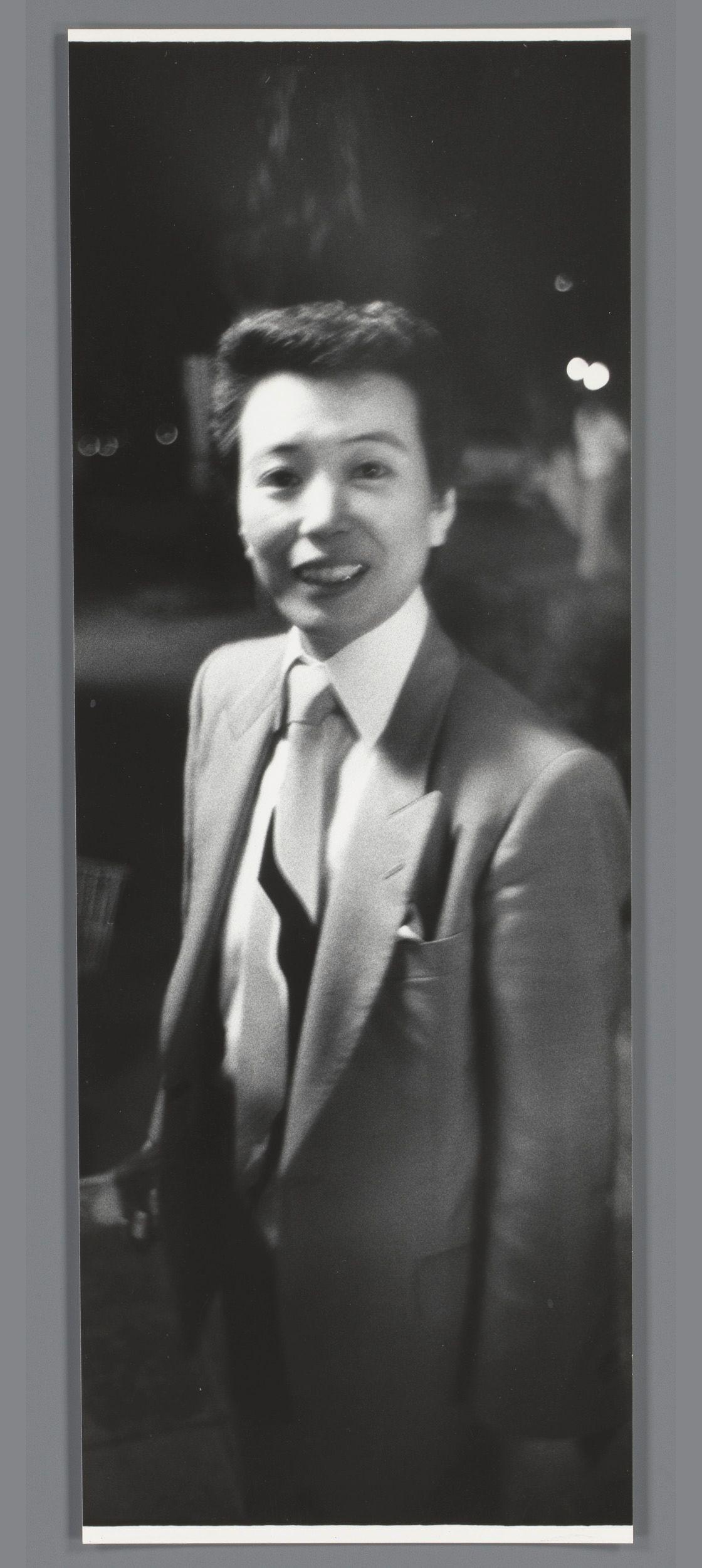 The image depicts a man standing in an indoor setting, possibly at night as suggested by the dark background. He is dressed formally with a light-colored suit jacket and trousers, wearing a white shirt and tie, along with a pocket square which adds to his polished appearance. His hair is styled upwards in what appears to be the 1950s fashion, suggesting that this photograph might date from around that era or represent someone who prefers classic styles. The man smiles broadly at the camera, showing teeth and eyes crinkled into an expression of happiness or amusement. There are bokeh lights visible on the left side in the background which hint at a bustling environment behind him. This gives context to his surroundings without revealing specifics about where he is standing - perhaps inside a venue like a club or event hall with some ambient lighting from overhead fixtures and decorative elements reflecting off surfaces.