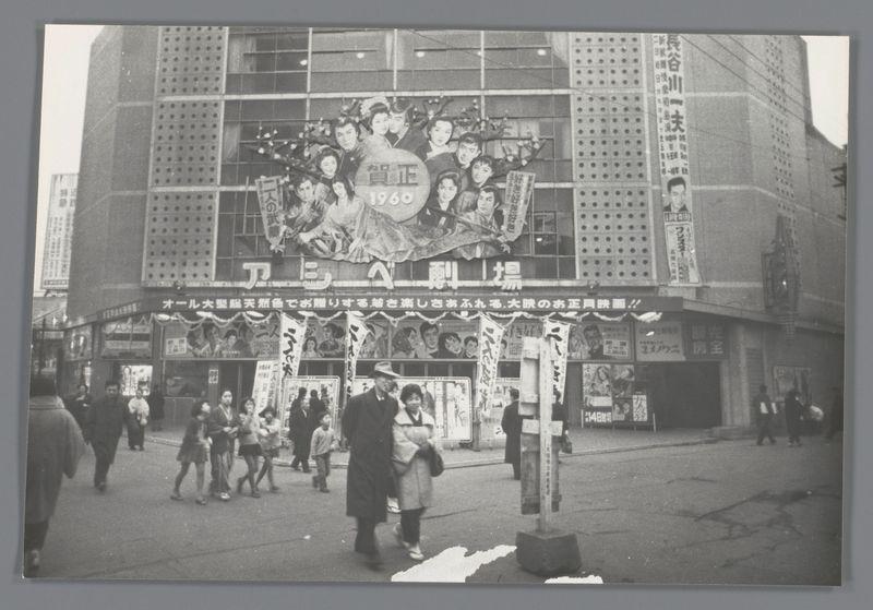 The image depicts a bustling urban street scene in Japan, possibly from the late 1950s to early 1960s. In the foreground, several individuals are walking or standing on a paved area that appears worn and cracked, indicative of high pedestrian traffic over time. The people vary in attire; some wear coats, while others have more casual clothing like skirts and jackets.

A large ornate banner with Japanese script stretches across the facade of an establishment at street level, likely advertising theater performances given its style and context within a densely populated urban environment. Above this building is another decorative sign featuring multiple individuals, possibly actors or characters from popular media, suggesting that it could be related to entertainment venues such as cinemas.

Additional signage includes what appears to be banners with Japanese text hanging above the street level, potentially indicating shops, theaters, or other commercial establishments. The buildings 