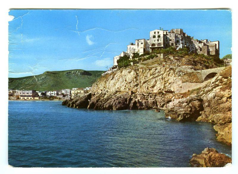 The image displays a scenic coastal town perched atop a rocky outcrop, overlooking the sea. The architecture of the buildings is Mediterranean, characterized by their whitewashed walls and terracotta roof tiles, indicative of historical or traditional construction methods often found in Southern Europe. These structures are layered on one another in an organic manner that follows the natural contours of the cliff face.

In contrast to this built-up area, a lush green landscape extends across the background, suggesting hills or mountains further inland which rise against the clear blue sky. The sea is calm with gentle waves lapping at what appears to be a rocky shore below and near where a car can be seen driving on a winding road that hugs the cliff edge.

A bridge crosses over part of this rugged terrain in the distance, emphasizing both human ingenuity and the natural barrier it has been built against. The overall atmosphere is one of tranquility and picturesque beauty, typical f