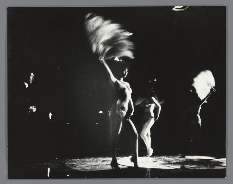 The image is a black and white photograph capturing a dynamic moment on stage. The main subject appears to be two performers, likely dancers or theatrical actors engaged in an energetic performance. One figure is captured mid-leap with their limbs extended dramatically, suggesting motion and perhaps part of a dance routine. Their hair seems caught in the wind, adding to the sense of movement.

The second performer stands near them, possibly interacting or preparing for their own move within the choreography. They are partially obscured by shadows and appear less focused than the central figure, which draws attention away from them but still contributes to the overall scene's atmosphere.

In addition to the performers on stage, there is a person off to one side of the image who seems to be part of an audience or perhaps another performer waiting for their turn. This individual stands out due to being in relative darkness compared with the illuminated figures and adds depth by indica