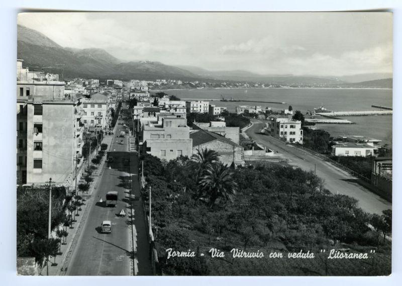 This black and white historical photograph depicts a panoramic view of Formia, Italy. The image captures the bustling street life in what appears to be an earlier time period, likely mid-20th century based on vehicle models present. On the left side is a multi-story building with visible signs and posters that suggest commercial use at its ground level.

The road runs through the center of the photo, flanked by trees and lined with various buildings including residential apartments to offices. The street is active with both pedestrians and vehicular traffic which includes cars indicative of post-war era design as well as a bus or public transport vehicle visible in one segment of the roadway. 

Moving towards the right side, there's an expanse that shows a clear delineation between urban architecture near water bodies and more natural landscape leading to mountainous terrain further back. The coastline is prominently featured with what appears to be port facilities extending into c