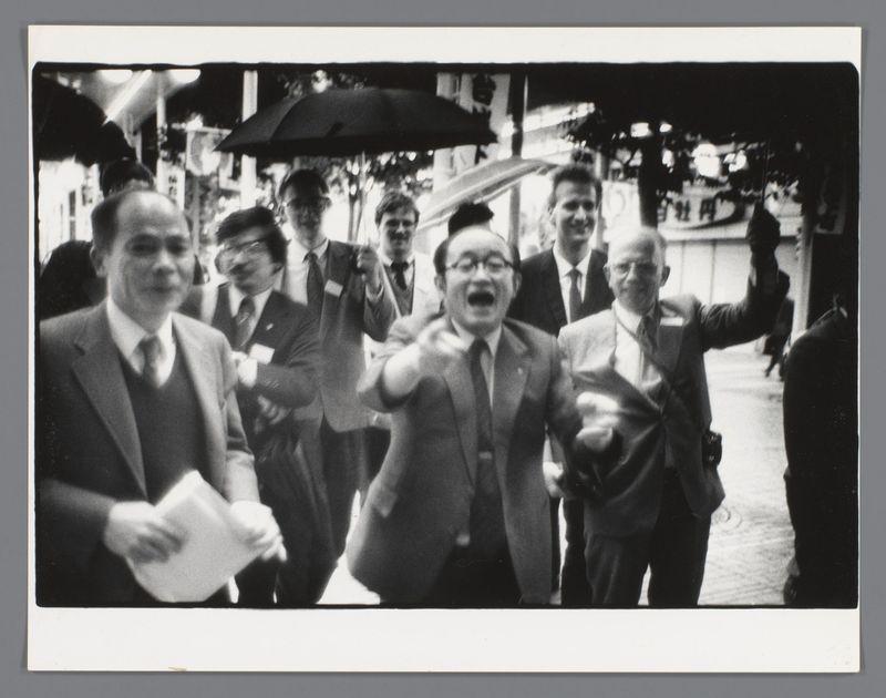 The image is a black and white photograph depicting several individuals walking down what appears to be a street or public area. The focus of the photo centers on five men in suits, with one man prominently positioned at the center appearing animated, perhaps speaking vocally while gesturing with his hands. He has glasses and wears a suit without a tie; he is flanked by two other individuals who are looking towards him, suggesting that they might be part of the same group or discussion.

To the left of this central figure stands another man in a coat holding what looks like some papers or documents while observing the situation with his gaze directed away from the camera. On either side of the center individual stand men wearing suits and ties; their expressions range from attentive to somewhat amused, indicating that they may be part of an ongoing event.

In the background, other figures can be seen walking by, adding a sense of movement and life to the scene. There's also visible
