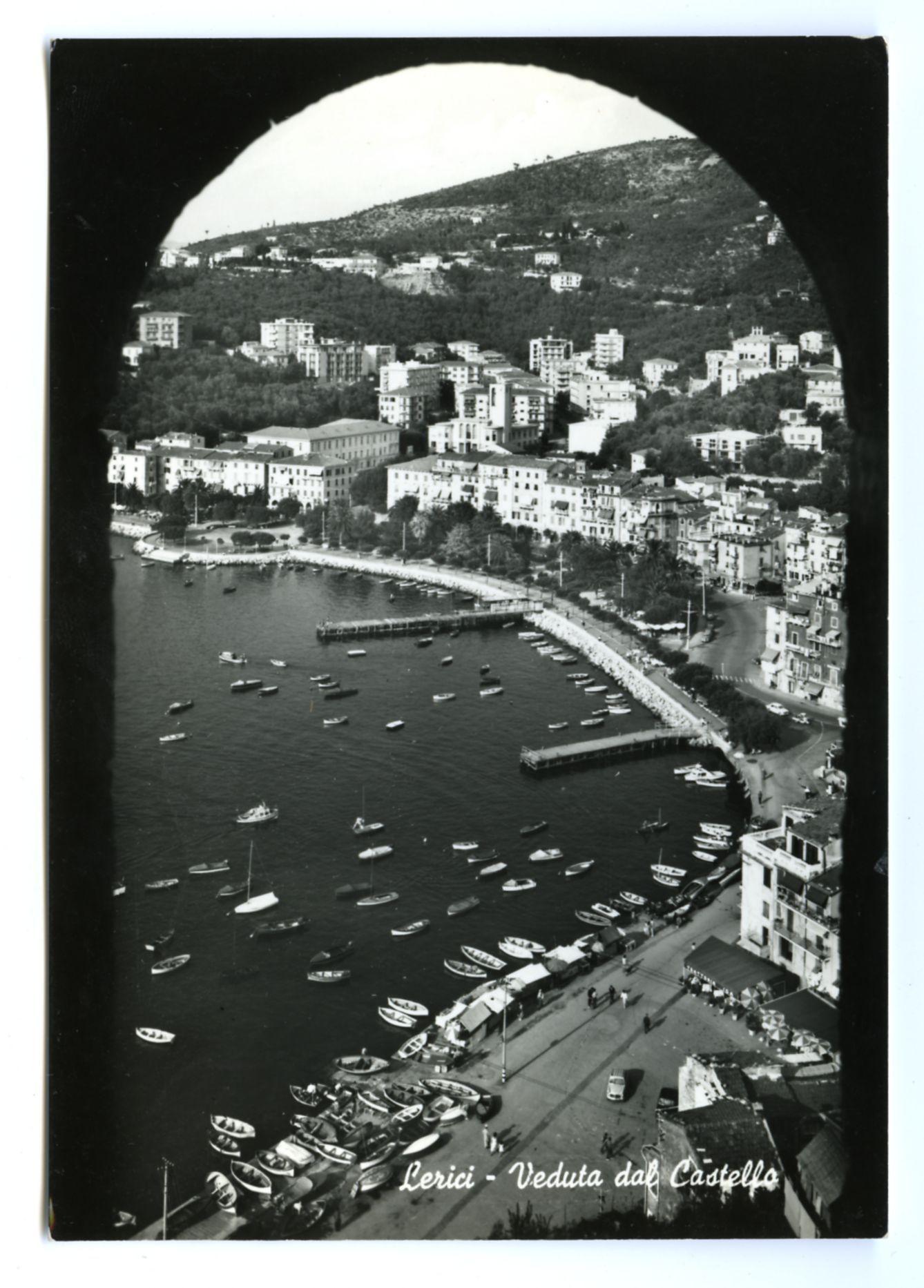 The black and white photograph captures a panoramic view of the coastal town of Lerici, with its dense cluster of buildings situated on rolling hills that descend to meet the sea. In the foreground is a harbor bustling with boats moored along piers extending into calm waters dotted with numerous small vessels. The water reflects the light differently than it does in direct sunlight, giving an almost ethereal quality to some parts while highlighting others. A road runs parallel to one of the piers where people can be seen moving about and umbrellas are set up alongside a market or cafe area near what appears to be a waterfront promenade.

Intriguingly, the view is framed by what looks like an archway or a natural opening in a hillside above. This suggests that the photo may have been taken from within another structure overlooking Lerici's harbor and town center. The perspective offers depth to the scene, with buildings receding into the distance up the hills.

Overlaying this histo