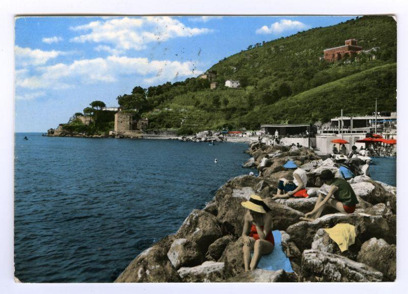 The photograph depicts a coastal scene with various individuals lounging on rocky outcrops by the sea. The sky is partly cloudy, suggesting it's likely late spring or summer. In the background, there are buildings nestled into green hills, and people can be seen near umbrellas at what appears to be an outdoor restaurant or cafe area adjacent to a small harbor where boats moored nearby indicate leisurely activity.