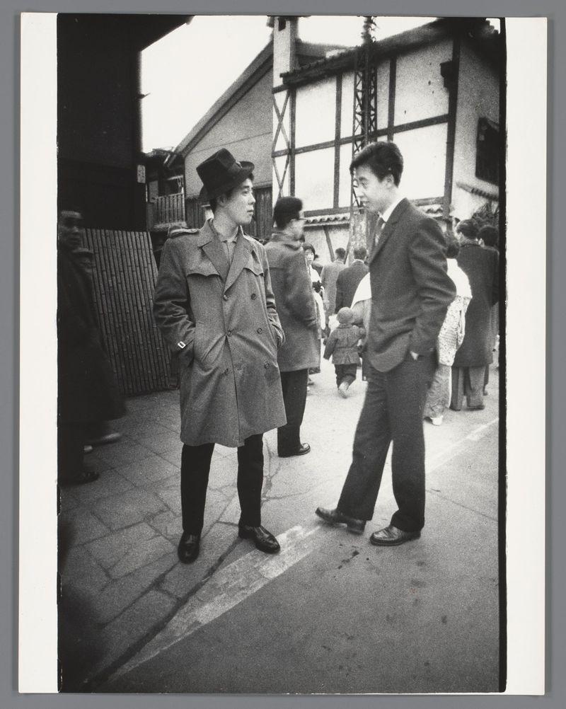 The image is a black and white photograph depicting two men engaged in conversation on a street. The man on the left wears a trench coat, dark trousers, and shiny shoes; his attire suggests practicality with an element of fashion from mid-20th century menswear. He sports a distinctive hat that resembles a bowler or derby style. His posture is relaxed as he turns slightly towards the other man.

The second man on the right stands in profile view facing away from us, wearing dark trousers and what appears to be a suit jacket with a lighter shade of material at his collar which could indicate an undergarment like a shirt. He wears polished shoes that are typical for formal wear during this period.

Behind them is a bustling street scene; other individuals appear walking or standing in the background, some looking engaged while others seem focused on their paths. There's architectural context provided by buildings with traditional East Asian design elements such as sliding doors and ti