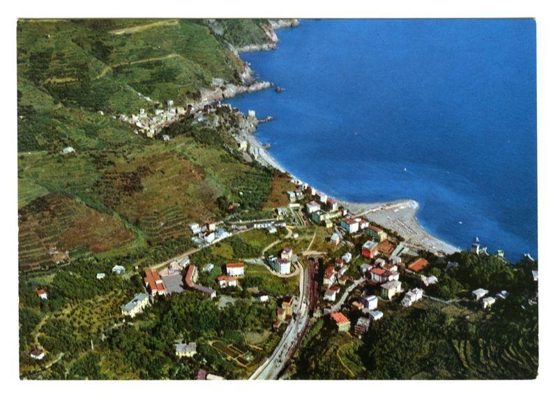 This image displays an aerial view of a coastal town nestled between lush green hills and the vibrant blue sea. The settlement is characterized by tightly packed, colorful buildings with red-tiled roofs that contrast against the verdant landscape. A winding road snakes through the heart of the village, connecting various streets lined with residences and small structures. Farmland or gardens can be seen interspersed among the built-up areas, indicating agricultural activity within this picturesque community.

The coastline features a sandy beach where people appear to be enjoying leisure activities near the water's edge. The sea is calm and vividly blue, extending into the horizon, and there are several boats scattered across its surface. On the right side of the image, rocky outcrops jut from the ocean towards the shore.

The overall composition captures a serene coastal community with natural beauty seamlessly blending human habitation within an idyllic setting.