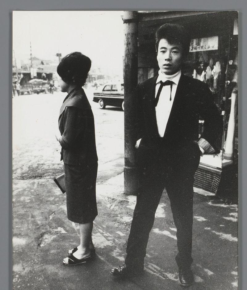 The image is a black and white photograph depicting two individuals on what appears to be an urban street. The person on the left stands with their body slightly turned towards the camera, looking away from it. They are wearing dark clothing: a short-sleeved jacket or coat over trousers, paired with sandals that have open toes. Their posture suggests they might be in mid-stride.

The individual on the right is facing directly into the camera and appears more formally dressed than their companion. This person wears a black suit with a white shirt underneath; no tie is visible. The style of the jacket sleeve indicates it's likely from an earlier era, possibly post-World War II based on fashion trends of that period.

Both individuals are standing in front of what seems to be a shop or establishment entrance, indicated by a door frame and hanging garments inside, which may suggest this photo was taken outside such a venue. In the background, we can see more people walking along with a