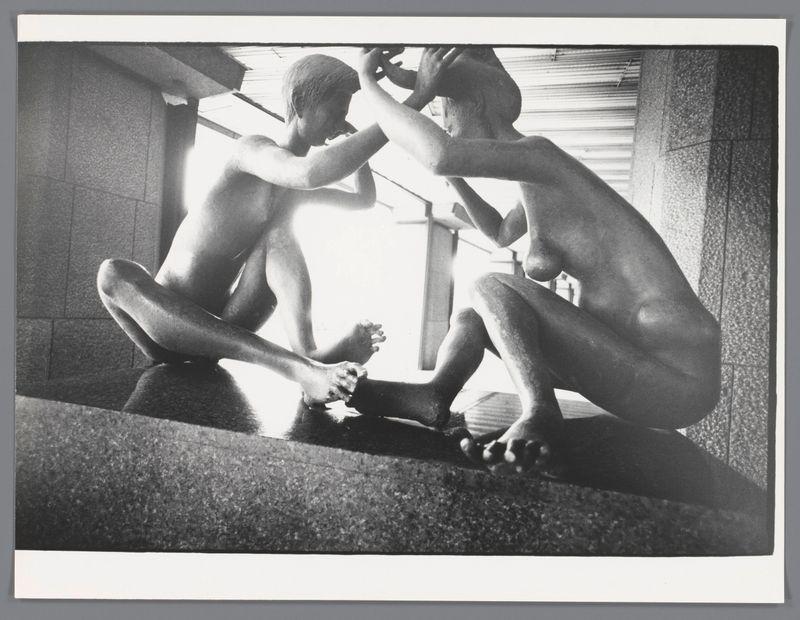 The image depicts a black and white photograph featuring two shirtless individuals, seemingly children or teenagers, engaged in an activity involving their feet. They are seated on what appears to be the edge of a bench or elevated structure with rough-textured surfaces. One person is squatting down, interacting closely with the other's leg while looking intently at something near his knee that cannot be clearly discerned from this perspective.

The scene takes place in an urban environment suggested by architectural elements such as tiled walls and possibly concrete structures around them. The lighting highlights their muscular forms and creates strong contrasts of light and shadow on their bodies, adding a dramatic effect to the composition.

In terms of context, it is noted that additional information about this image can be found through reference links provided in the original description source (https://images.loener.nl/EdvdElsken/full/6484/64844a46ba25398d90834800.jpg). This