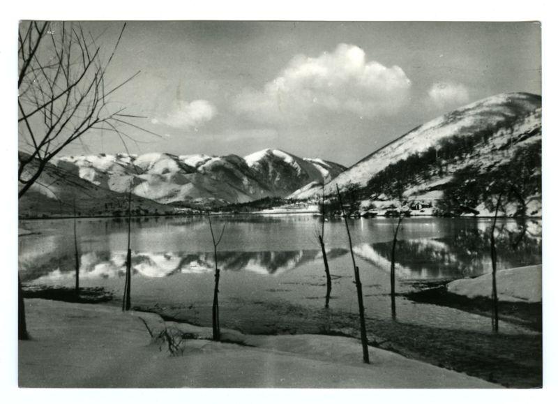 The image is a black and white photograph depicting a serene winter landscape with snow-capped mountains, barren trees, and calm waters reflecting the surrounding scenery. The sky has patches of clouds, indicating it might be midday or afternoon on an overcast day. There are several leafless tree branches in the foreground that frame the view into the lake, which appears still enough to mirror the mountainous background perfectly.

In the distance, a small cluster of buildings is visible near the base of the mountains, possibly suggesting a remote village or settlement nestled between the towering peaks and calm waters. The overall atmosphere conveyed by this photograph exudes tranquility and isolation from urban life, inviting contemplation on nature's quiet beauty during winter months.