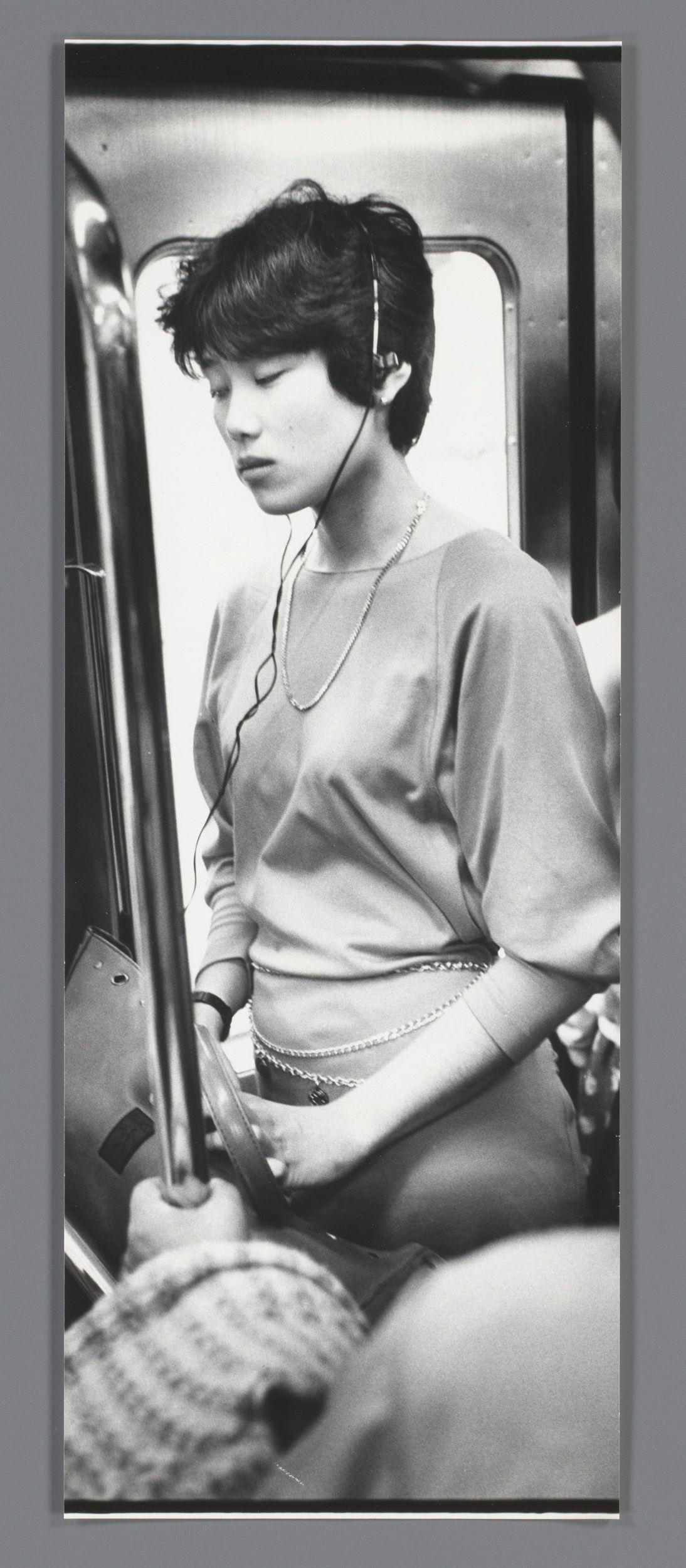 The image is a black and white photograph depicting an individual, presumed female due to the hairstyle and jewelry. The person appears seated in public transportation, likely on a bus or train given the visible seat poles typical of such vehicles. She wears headphones connected by wires that lead out of view, suggesting she may be listening to music or another audio source. Her attire consists of what looks like a light-colored blouse with long sleeves and is adorned with layered necklaces adding an element of sophistication to her outfit. The individual's expression is neutral, focused on something outside the frame which could indicate deep concentration or introspection as they gaze away from the camera lens. There are no other passengers in immediate proximity that can be seen within this narrow field of vision.