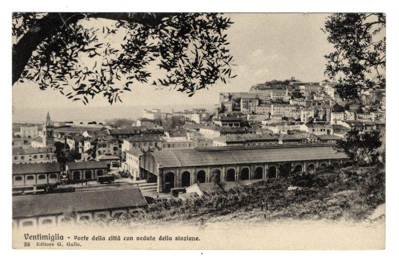 The image depicts a historical black and white photograph of the Ventimiglia Port. It features an elevated perspective overlooking what appears to be a bustling town with numerous buildings, including prominent structures such as a large covered market hall in the foreground that runs parallel along one side of a street or platform. The architecture suggests European influence, possibly Italian due to references like "Porte della città con veduta della stazione," which translates from Italian meaning “Doors of the city with view of the station.” A dense cluster of buildings is seen on elevated ground towards the right-hand part of the image and a church or tower-like structure visible in the middle distance, hinting at historical significance. The foreground shows natural elements like trees, indicating this may have been taken from an area near nature overlooking urban development.

A caption identifies "Ventimiglia" as both Italian text ("Porte della città con veduta della stazione