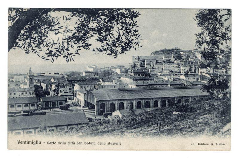 The image is a black and white vintage postcard depicting the Ventimiglia town, with specific focus on part of the city including views from around the train station. The photograph showcases an expansive view of urban architecture nestled among hills, with multiple buildings ranging in height but generally low to mid-rise structures typical for European towns during that era.

Prominent features include a large covered structure resembling a railway waiting hall or goods shed at the bottom left corner and another similar building further back towards the center. The town is characterized by its densely packed housing, terracotta roofs indicative of Mediterranean architecture, narrow streets winding through the area, and various small buildings possibly shops or residences.

The upper portion of the photograph captures an expansive view over rooftops with a tree branch partially framing the top edge on both sides. In the far background, there's what appears to be fortification or h