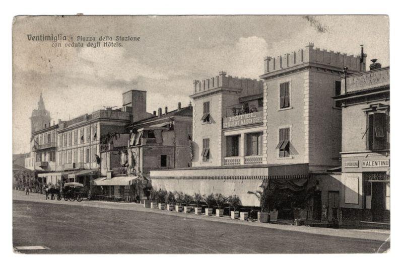 The image depicts a historical black and white photograph of the Piazza della Stazione in Ventimiglia, featuring buildings with European architectural styles. Prominent structures include "HOTEL Maison Doree" on the right side and an old-fashioned vehicle near the left sidewalk. The scene appears calm without any visible people or modern elements, suggesting a moment captured from the past. This image is used as part of historical documentation for FerroCandilera's collection, referenced by their webpage link in the alt text provided above this description.