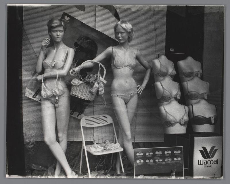 The image depicts a vintage-style photograph of two mannequins dressed in lingerie displayed against what appears to be the interior wall or storefront. The left side shows a full-body view of a female mannequin wearing brassiere and panties, holding a basket containing undergarments on its arm while seated next to an open chair with a pair of sandals beside it. A second headless upper torso is visible in front of this figure.

The right features another display featuring multiple bust forms with bras fitted onto them; these displays are arranged vertically. To the far left, there's partial text that includes "Wacoal," suggesting the brand or store name associated with this lingerie display.

The overall setting has a monochromatic color scheme and appears to be an advertisement for Wacoal lingerie. It seems like it could have been captured during a time when such displays were more common in retail environments, possibly from around 1975-1990 as indicated by the reference informat