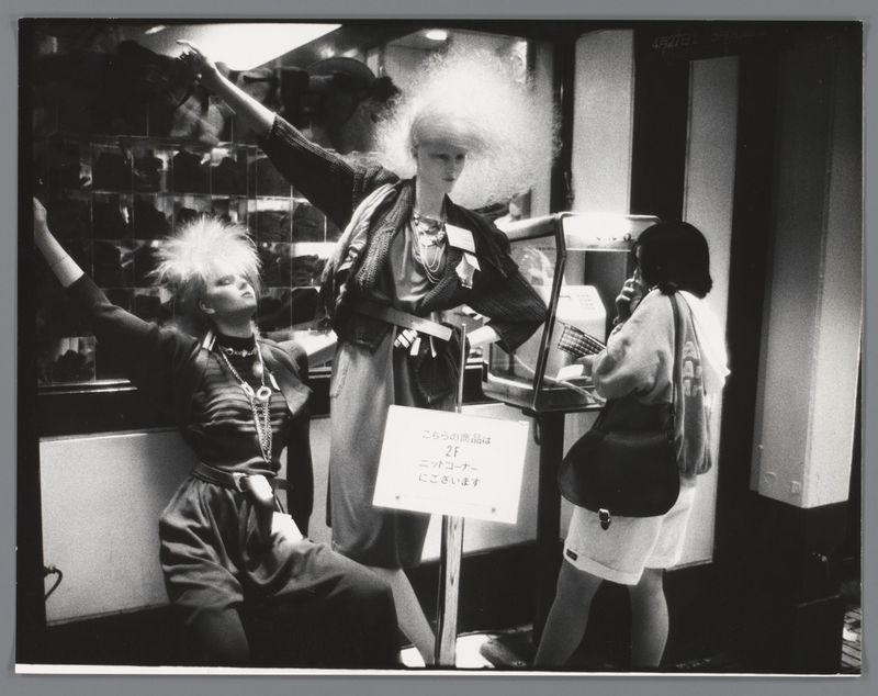 The image captures a black and white street scene featuring three individuals. The central figure is standing, wearing an elaborate outfit with layers of clothing, accessories like necklaces and bracelets, and holding what appears to be money or cards in one hand while reaching out towards the second individual on the right side. This person, dressed casually in shorts and a t-shirt, holds a purse over their shoulder and seems engaged in conversation via a mobile phone.

On the left is another figure seated with an arm raised, wearing clothing that suggests casual style but also has distinctive elements like stripes or patterns. They have short hair and are adorned with jewelry including rings on fingers extended toward the standing individual. 

A signboard stands prominently between these two individuals; it seems to indicate a queue system for entry into a location. The background is indistinct, suggesting an urban environment with various lights and reflections hinting at comme