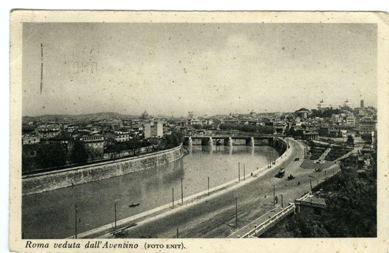 The image depicts a historical black and white photograph of Rome, captured from the vantage point known as Aventino. It showcases an expansive view of the Tiber River winding through the cityscape with bridges crossing over it. The riverbanks are lined with streets featuring tram tracks or roads that have very few vehicles on them, indicating this could be a scene from mid-20th century urban life in Rome.

In the background, various buildings and structures can be seen, including what appears to be governmental edifices along the left bank of the Tiber. The horizon shows rolling hills with more construction or possibly other cityscapes further away under an overcast sky. There are no people visible in this scene; instead, it focuses on urban architecture and infrastructure.

The photograph carries a caption that reads "Roma veduta dall'Aventino (FOTO ENIT)," which translates from Italian to English as "View of Rome taken from Aventino." This indicates the image was professionally 