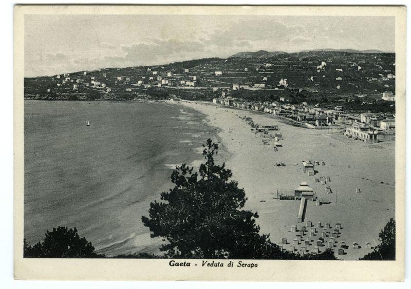 The black and white photograph showcases a panoramic view of the beachside town Gaeta, known as "Veduta di Serapo" in Italian. The image is taken from an elevated vantage point overlooking a coastline where a sandy beach stretches into calm waters with a few small boats dotting the horizon line. There's evidence of early 20th-century architecture and infrastructure along the coast, including rows of wooden sunshades or shelters, possibly used for bathing facilities.

The town rises gently on a hillside above the sea, densely packed with buildings that hint at residential areas and perhaps some commercial establishments near the shore. In the distance, more hills form part of the landscape's backdrop, creating a layered effect against an overcast sky which suggests either early morning or late afternoon light conditions. A lone sailboat is visible on the water, adding to the serene atmosphere.

In terms of historical context, such postcards were popular mementos for travelers in tha