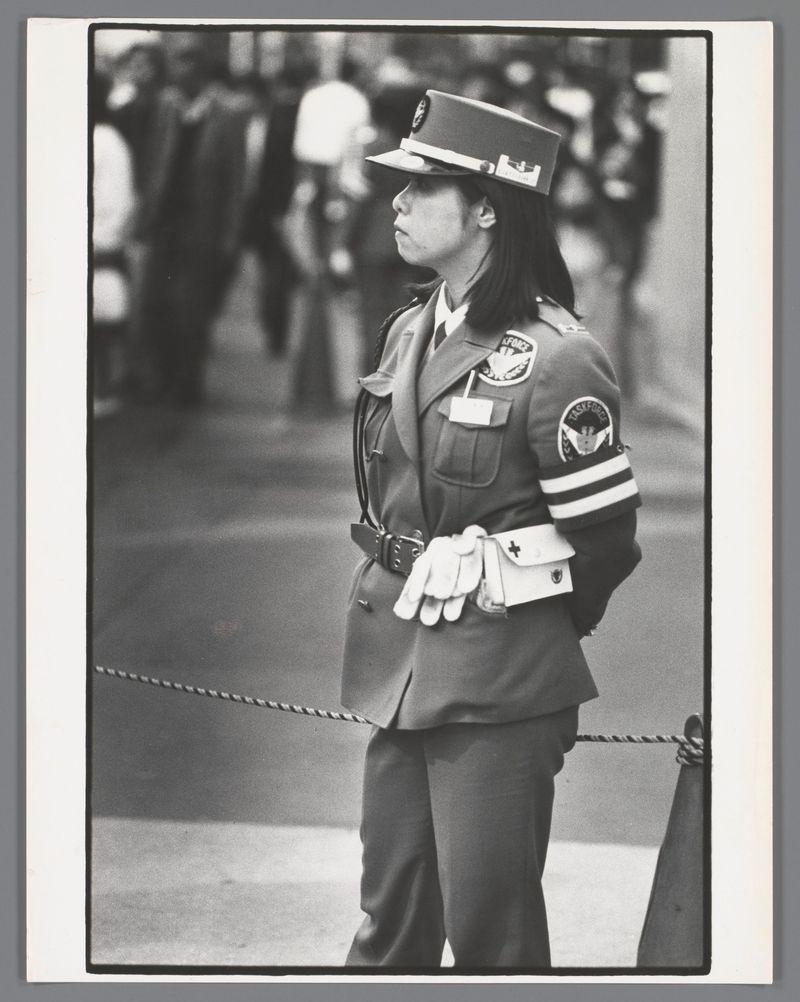The image depicts a woman in uniform standing on guard duty. She is wearing a cap with insignia, suggesting she may be part of a security or police force. Her jacket has patches and badges indicating her affiliation to an organization labeled "Task Force" alongside other text that isn't clearly legible due to the resolution. In one pocket of her jacket, there's what appears to be gloves typically used in medical or paramedic services, with visible crosses on them which are commonly associated with first aid service uniforms.

She stands upright and looks off into a distance over the rope barrier she is positioned next to, possibly indicating control over an entrance point. The background shows out-of-focus figures of people walking by, implying this scene takes place in a public space or event where security measures were likely implemented due to crowd presence. There's no clear indication as to whether it’s indoors or outdoors because the lighting doesn’t provide enough cues.

Th