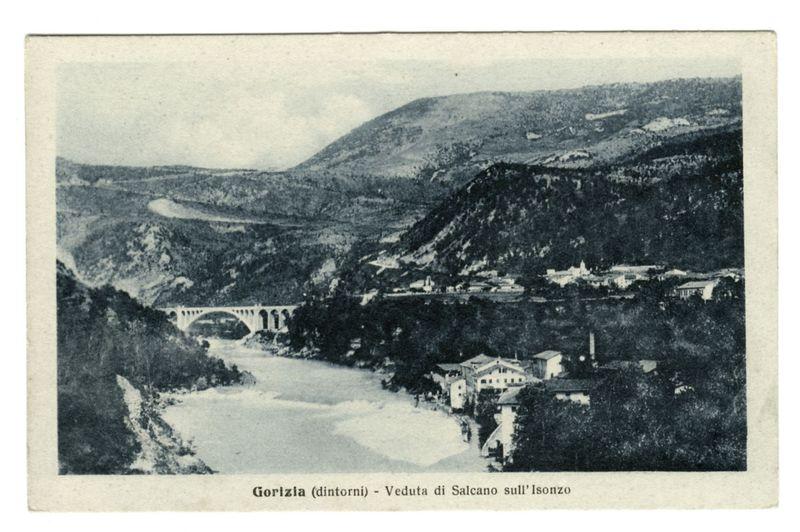 The image is a black and white vintage postcard depicting the landscape around Gorizia, specifically highlighting "Veduta di Salcano sull'Isonzo" which translates to 'View of Salcano on the Isonzo'. The scene shows a river winding through hilly terrain with lush vegetation. A prominent feature in the picture is an arched bridge spanning across the water at one point. In the midground, there's a cluster of buildings and structures nestled among trees. Notably, this image was sourced from FerroCandilera based on their watermark or reference information provided alongside another URL which leads to more details about Gorizia (Dintorni).