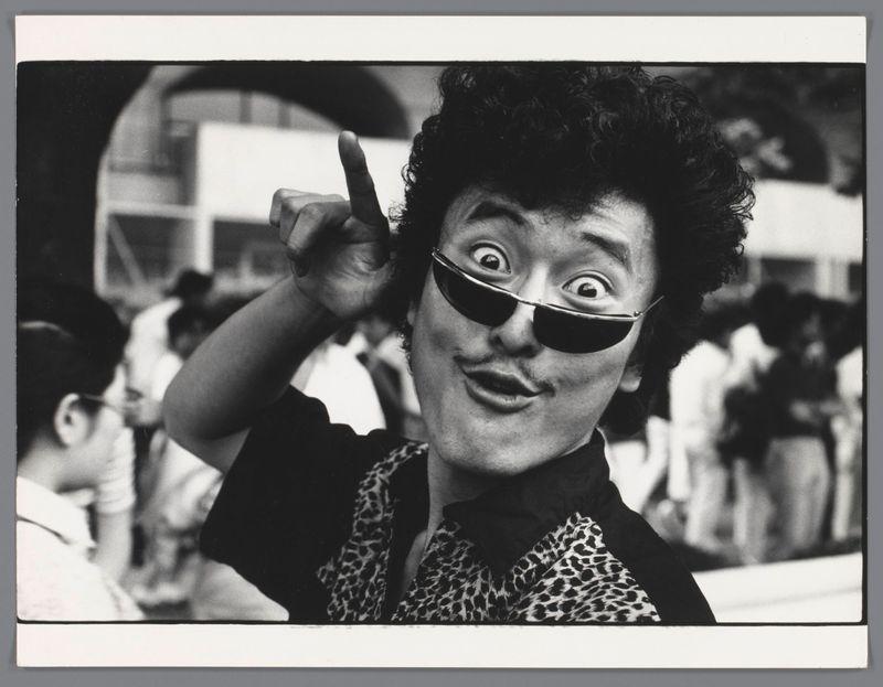 The image is a black and white photograph showing an individual with curly hair, wearing large-framed sunglasses. The person appears to be in motion or caught mid-action as indicated by the blurred background suggesting movement. They have one hand raised near their head, pointing upwards with their index finger.

They are dressed in what seems like a leopard print garment which is partially visible and has a dark collar around the neck area. The backdrop behind them shows indistinct figures of people who appear to be at an event or gathering, as suggested by partial views of others' clothing styles that include varied patterns such as stripes on one figure.

There's no clear context provided about the location, but there are hints suggesting it might have been taken in a public place with multiple individuals around. The image has visible creases and wear marks indicative of an old photograph or printed material which could suggest its age or handling over time.