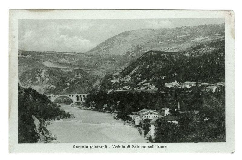 The image shows a vintage black and white photograph of Gorizia, identified as "dintorni" or surroundings in the caption. It features an elevated perspective overlooking a river flowing through hilly terrain with lush vegetation on both banks. A bridge spans across one portion of the river amidst greenery, suggesting it is part of Salcano sull'Isonzo (Salca Bridge). The landscape appears undisturbed and pristine. Traditional buildings are visible along the riverbanks near what seems to be a town or village situated within this scenic setting. Above these structures, on elevated terrain, there appear to be more residential areas with various houses and possibly small-scale industrial establishments scattered across rolling hills. In the distance, atop the hillside, is an area that might include public buildings such as churches or administrative centers due to their prominent placement in the landscape design of towns during historical periods. The sky above shows a faint cloud cover in