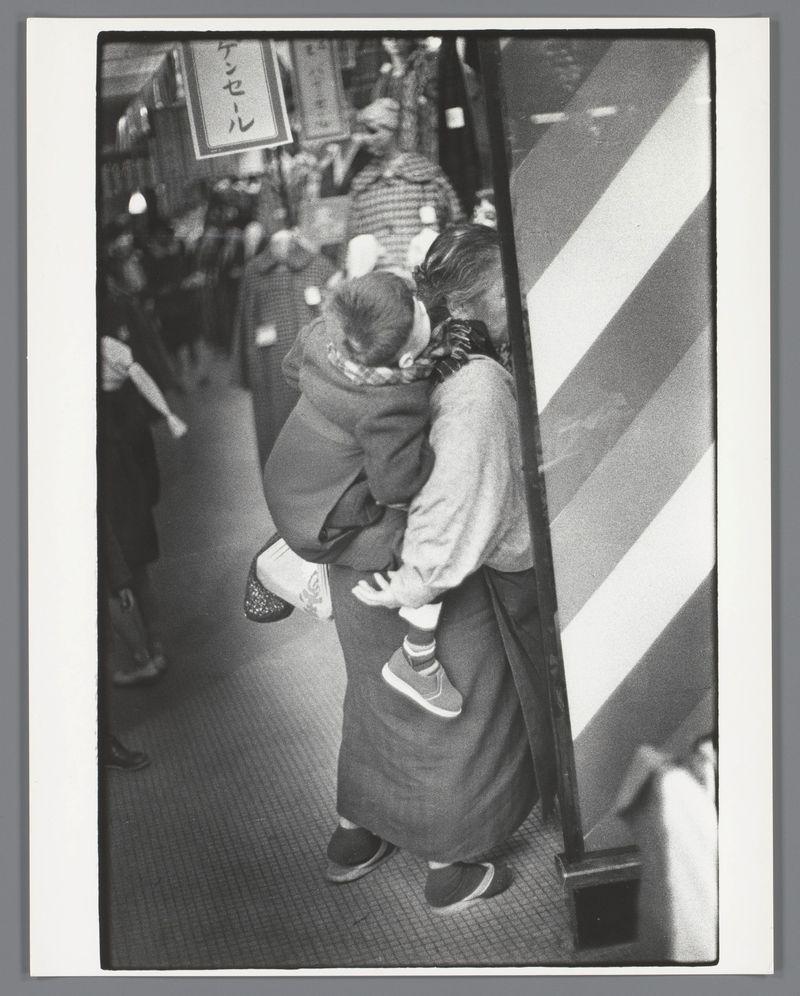 A black and white photograph depicting a woman carrying an infant on her back. They are in what appears to be a crowded urban street, with various pedestrians visible around them. The child is wearing a light-colored hat or head wrap, while the adult's attire includes dark pants and shoes paired with a long-sleeved garment that reaches down past their knees. Notably, they're both dressed for cool weather. A striped barrier runs parallel to the walkway where this duo travels. Japanese characters are visible in the background on an overhead signboard or storefront banner, indicating the setting is likely Japan during 1950s-60s due to fashion and language presence.
