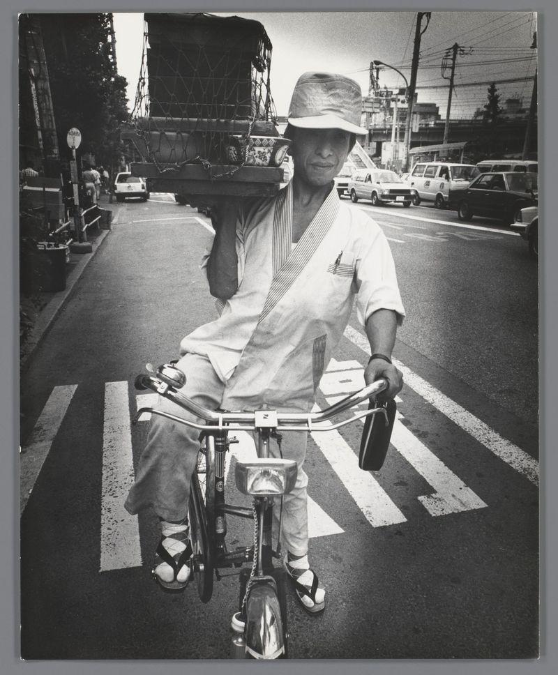 The image depicts an individual riding a bicycle on what appears to be a street or road. The person is wearing casual attire, including sandals and carrying several items in their hands while pedaling the bike. They are navigating through an urban environment with various vehicles visible around them, such as cars parked along the roadside.

Black-and-white photography adds a timeless quality to the image, emphasizing contrasts between light and shadow on textures like clothing folds or shadows cast by objects. The composition is framed within what seems to be a vintage border, which suggests that this photograph might have historical context.