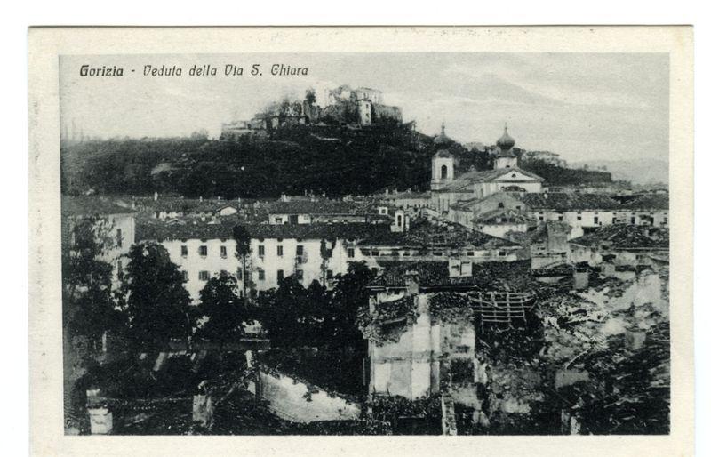 The image depicts a historical black and white photograph of the town Gorizia, with its prominent location on an elevated hill. The caption indicates that this is a view from Via S. Chiara. In the foreground, there's evidence of destruction or decay in what appears to be residential buildings, suggesting possible post-war damage or neglect. Several structures exhibit signs of disrepair such as missing roofs and exposed brickwork.

Dominating the background are multiple church spires with domed tops that stand out against a faint skyline, hinting at Gorizia’s historical architecture and possibly its religious significance. The vegetation on the hill adds texture to the upper portion of the image while providing contrast to the man-made structures below. Overall, the scene conveys a sense of history and perhaps resilience in the face of adversity.

The black-and-white format emphasizes contrasts between light and shadow, highlighting architectural features without color distraction. 