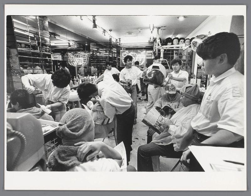 The image is a black and white photograph capturing the interior of what appears to be an old-fashioned barber shop. Multiple people, both standing and sitting, are present within this setting. The seated individuals seem mostly male with one notable female among them who stands out due to her haircut that looks like it could belong in the 1970s or early '80s.

The staff, possibly barbers given their attire resembling white aprons and rolled-up sleeves, is engaged variously: combing hair, holding mirrors, using scissors for cutting, talking amongst themselves, and interacting with customers. The shop itself has a dated look; the ceiling lights are round lamps typical of mid-century design, there's an abundance of reflective surfaces like large mirrors on walls, and shelves filled with bottles which might contain grooming products.

The environment is crowded yet orderly, suggesting it’s during peak hours when many patrons feel comfortable getting their hair done. The overall atmosp